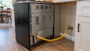 The back of a slide-in range pulled forward in a kitchen showing a yellow flexible gas connector and a shutoff valve